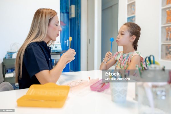 Female speech therapist working with elementary age girl, using special tool, speech therapy techniques