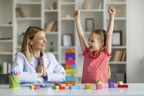 Cute Little Girl And Child Development Specialist Celebrating Therapy Progress During Meeting In Office, Smiling Therapist Lady Clapping Hands While Happy Female Kid Raising Fists And Shouting Hooray
